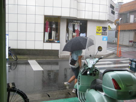 雨の中を歩く人と、店舗前に停められたベスパのある風景。