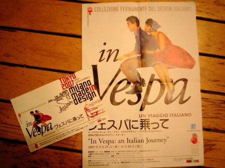 「in Vespa」展のポスターとチラシが木のテーブルの上に置かれている写真。