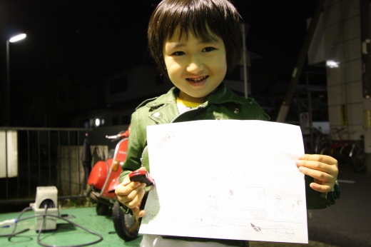 笑顔で自作の絵を見せる男の子と後ろに赤いベスパ