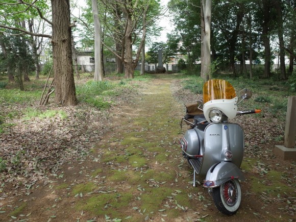 林の中の小道に停めた銀色のベスパ。奥には朽ちた建物が見える。