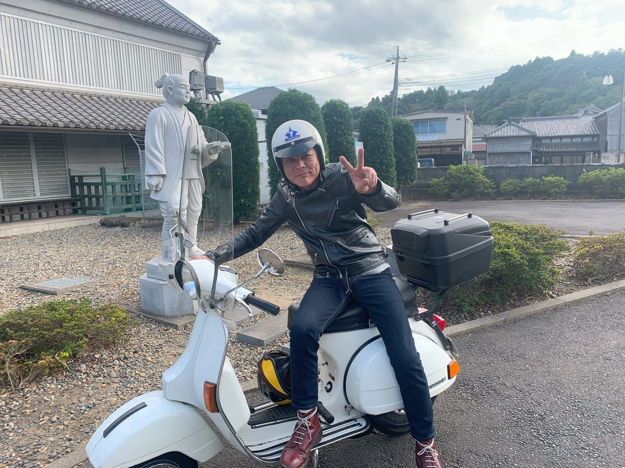 白いベスパにまたがりピースサインをするライダー。背後に伊能忠敬の石像が見える。