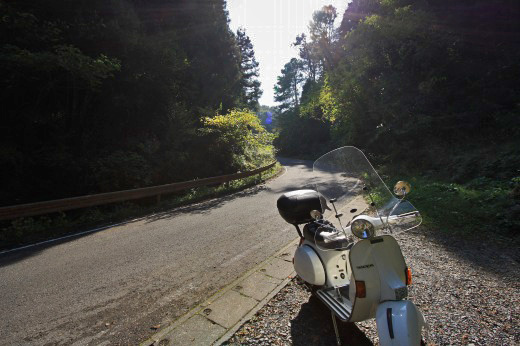 山間の静かな峠道に停めたベスパ。朝日が差し込み、木々の陰影が美しい。