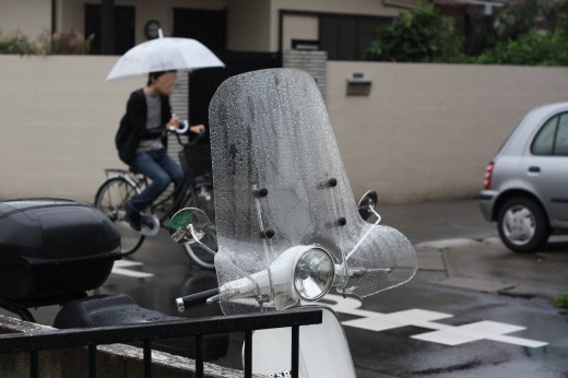 雨に濡れたベスパと風防、背景に傘の自転車