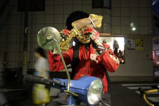スルメを両手で広げて顔を隠すベスパ乗りの男性（夜の街角）