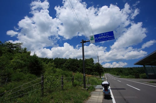 パノラマライン入口を示す標識とベスパ。夏空に浮かぶ雲が印象的な高原の風景。