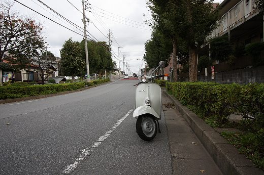畑のあぜ道に停車した黄色いベスパ、ミラー越しに広がる曇り空と街並み。