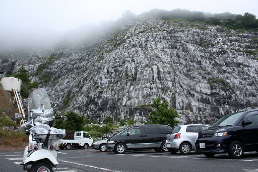 岩肌が迫る駐車場に停まる白いベスパ。霧がかかった幻想的な風景。