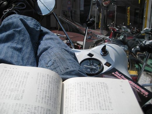 日向に停めたベスパの上で足を伸ばしながら読書しているリラックスした光景