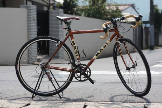 茶色のMASIシクロクロスバイクが街中でスタンドに立つ様子
