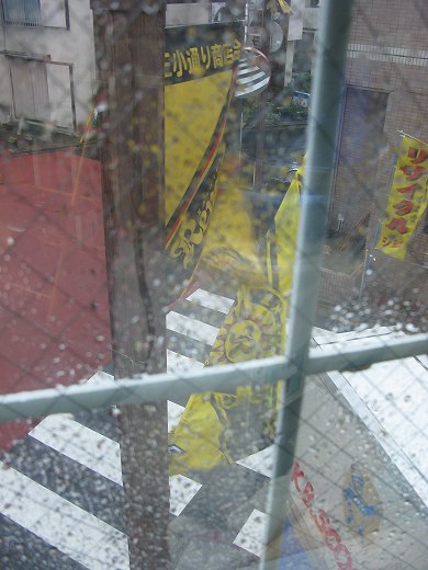 雨に濡れた窓越しに見えるK.B.SCOOTERS店頭の黄色い旗と交差点