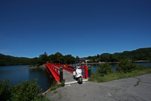 湖上に架かる真っ赤な橋とベスパ。深い青空と湖面のコントラストが美しい一枚。