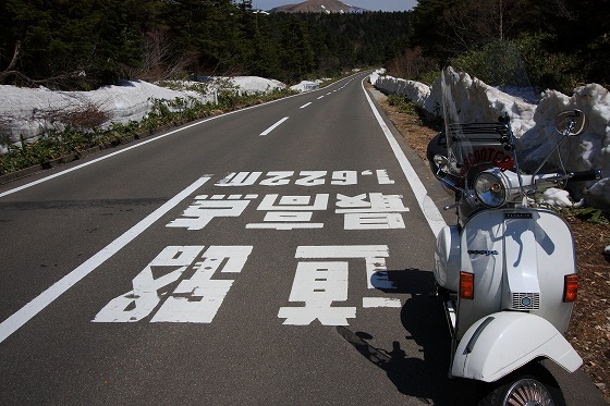 磐梯吾妻スカイラインの道路に描かれた標高表示と、残雪の中を走る白いベスパ