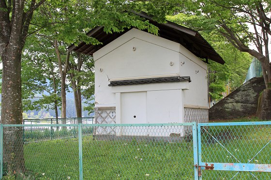 木々に囲まれた白壁の土蔵と、手前のフェンスが並ぶ風景