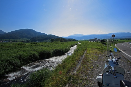 小川沿いの土手に停めたベスパと、遠くに続く山並み。のどかな田舎道の夏景色。