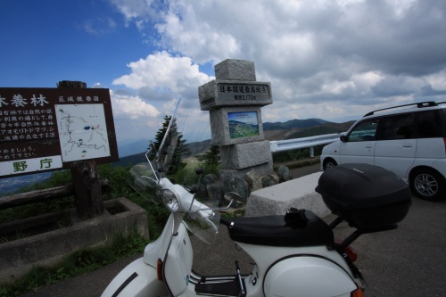 日本国道最高地点の標識とベスパ。雲が広がる山岳パスの絶景スポット。