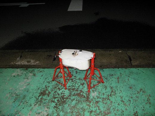 夜間、路肩に置かれた白いフューエルタンク。赤い台に乗せられている。背景は濡れた道路面。