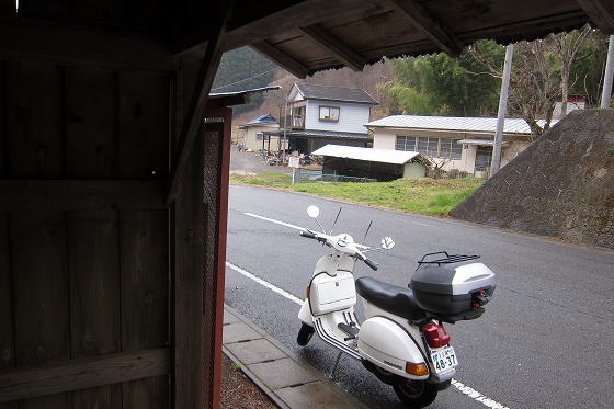 田舎の屋根付きバス停で雨宿りする白いベスパ
