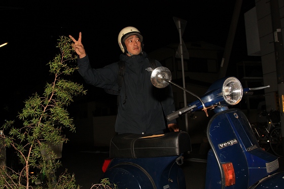 ギターを背負ったSさんがバイクの横で笑顔を見せている様子