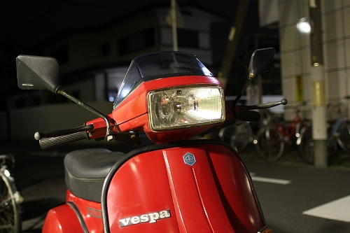 夜の街角に停められた赤いVespa PX125T5、ヘッドライトが灯っている。