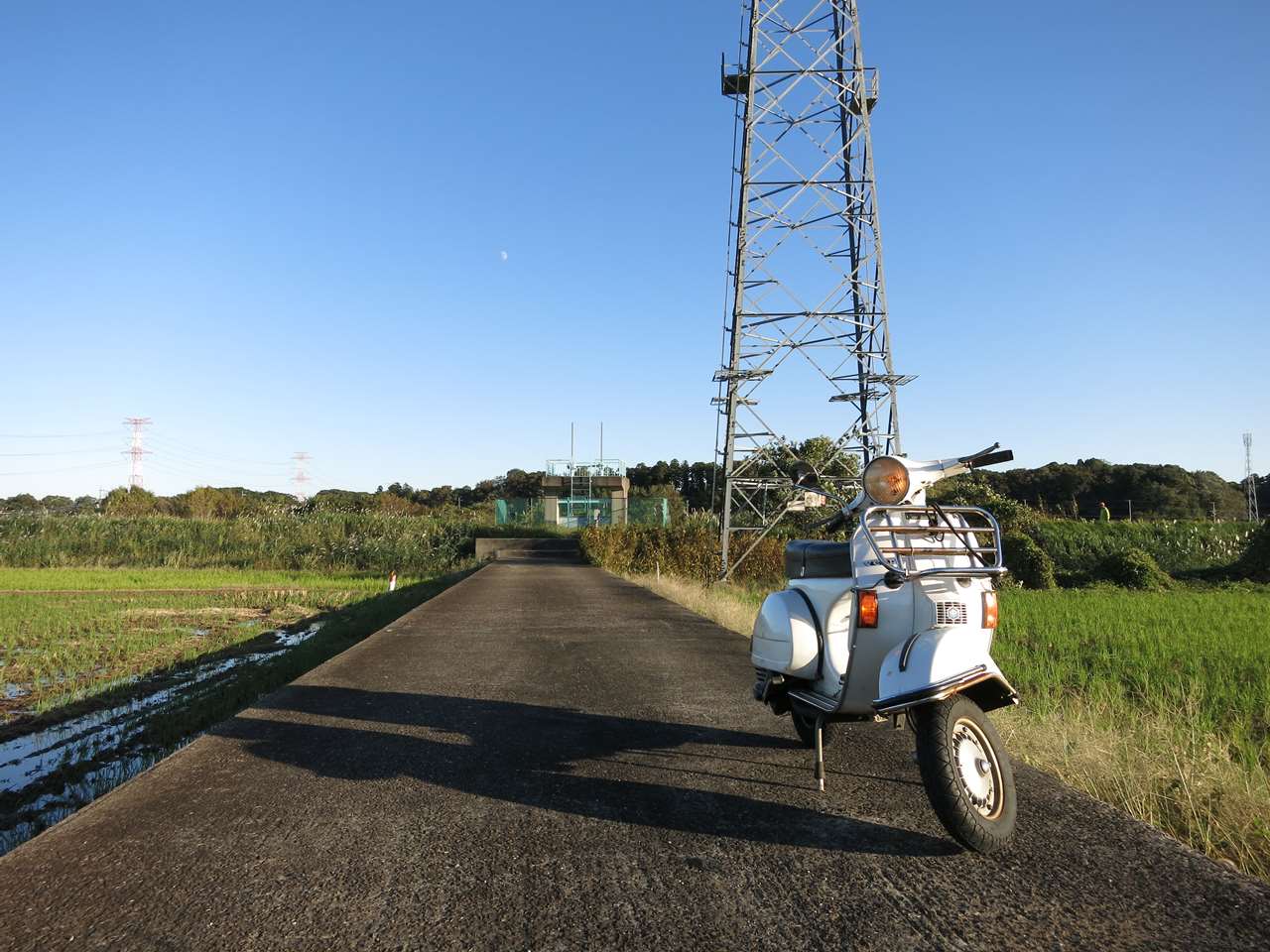 水田沿いの舗装路に停まっているベスパP150Xの試運転風景。背景に電波塔と青空が広がる写真