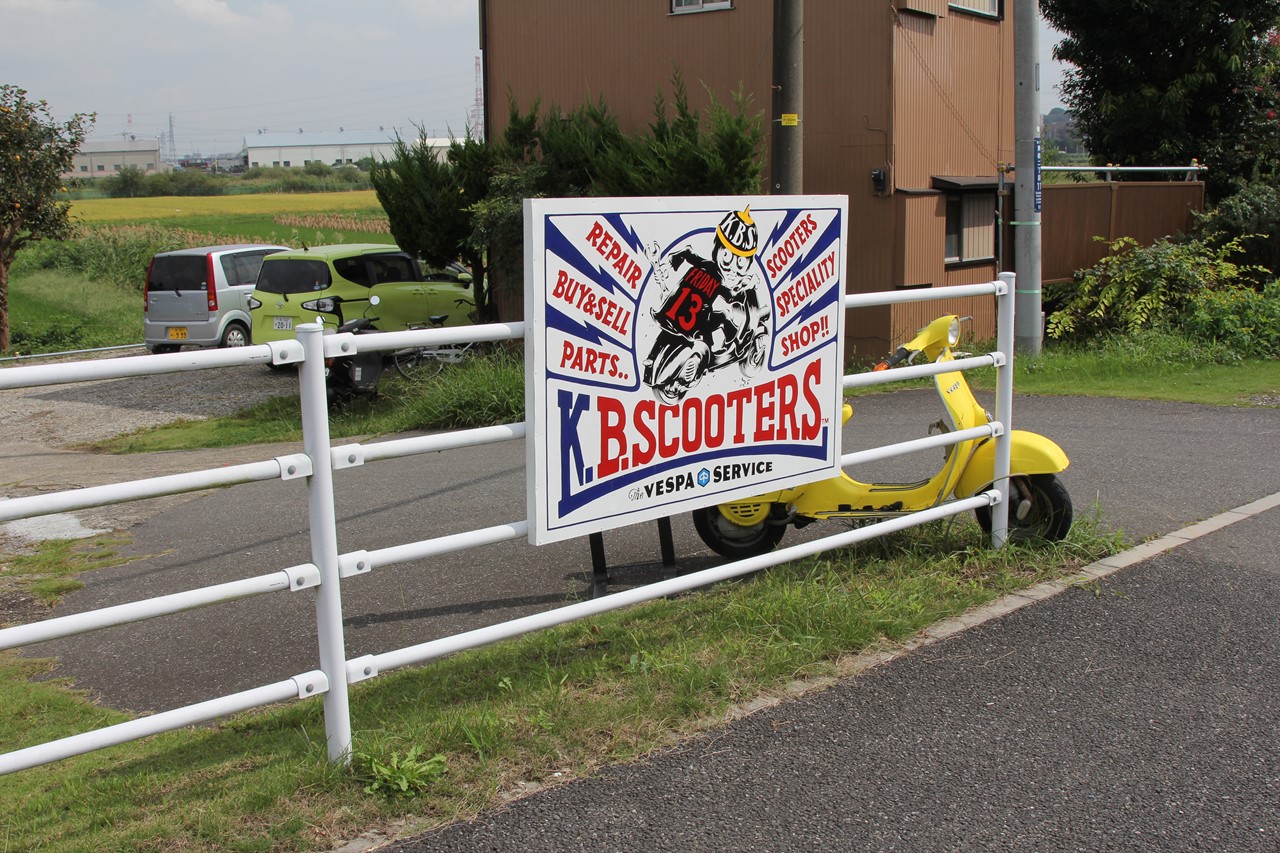 K.B.SCOOTERSのショップカードと点火プラグ、背景は屋外の風景